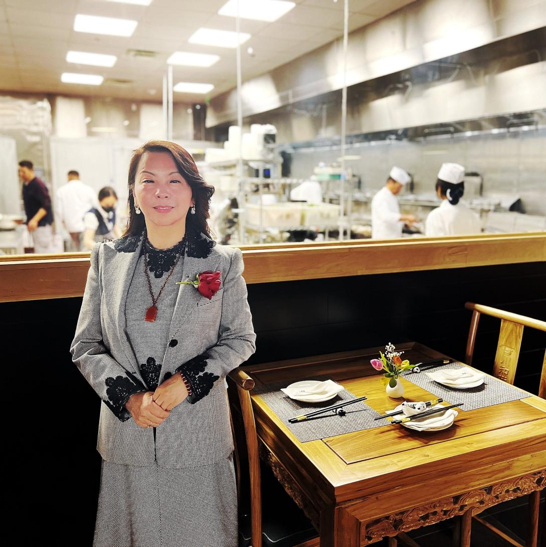 Owner Ping Wu standing next to a table at the restaurant, with the kitchen full of chefs working behind her.
